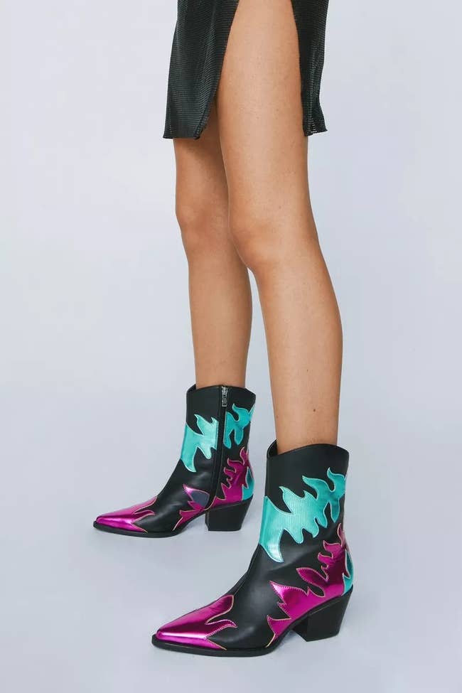 model wearing black pointy toe low heel black ankle boots with hot pink and teal metallic details and a side zip fastening