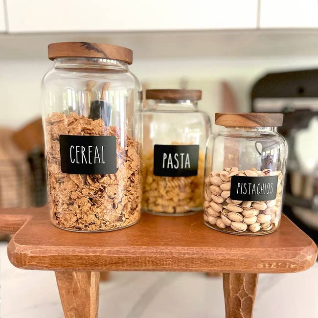 Three labeled glass jars containing cereal, pasta, and pistachios on a wooden shelf