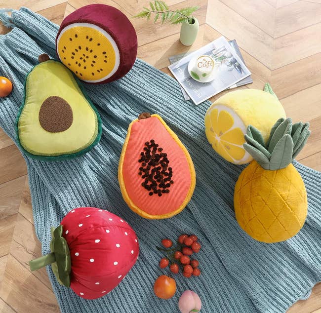 Plush effect   pillows, including avocado, papaya, pineapple, lemon, passionateness  fruit, and strawberry, arranged connected  a bluish  knitted blanket