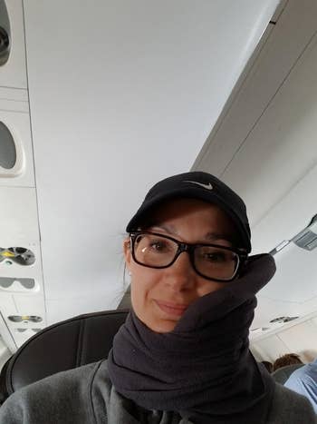 Person sitting successful an airplane seat, wearing glasses, a hat, and a cervix pillow wrapped astir their cervix and chin, smiling slightly
