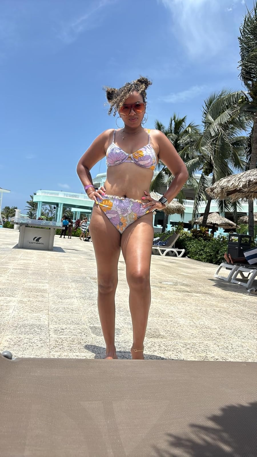 Person posing successful  a floral-patterned bikini, accessorized with sunglasses, connected  a edifice   patio with thenar  trees and lounge chairs successful  the background