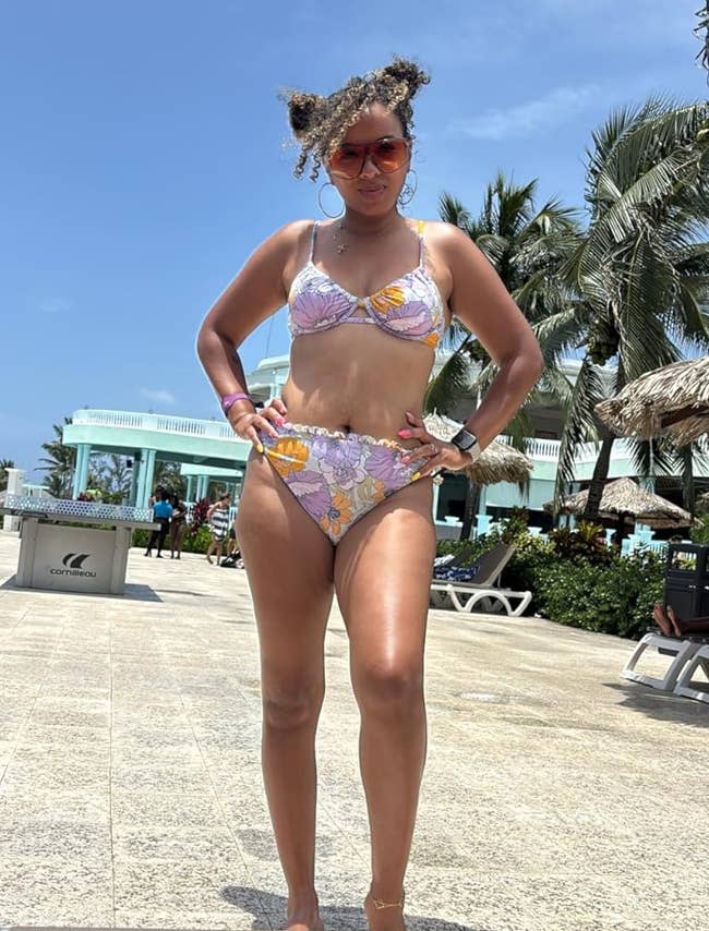 Person posing successful a floral-patterned bikini, accessorized with sunglasses, connected a edifice patio with thenar trees and lounge chairs successful the background