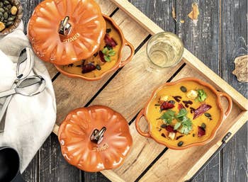 Two pumpkin-shaped orangish Staub bowls with lids connected a woody tray, containing crockery garnished with herbs and seeds, adjacent to a solid of achromatic wine