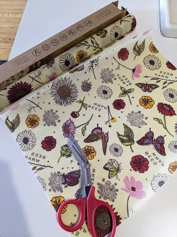 Beeswax wrap with floral and butterfly design is being cut with scissors on a kitchen countertop. Eco-friendly alternative to plastic wrap