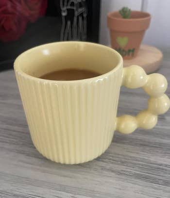 Yellow ribbed mug with a unique bead-like handle filled with coffee, placed on a wooden table near a potted plant and picture frame