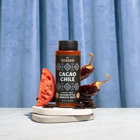 Bottle of Tenavo Cacao Chile sauce with tomato slice, chili peppers, and chocolate pieces arranged artistically on a light surface