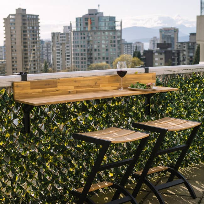 Outdoor balcony bar with wooden counter, two stools, a glass of red wine, and a small plate of food with a city skyline in the background