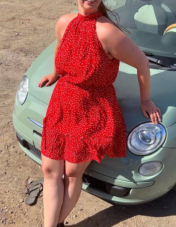 reviewer in a sleeveless red polka-dot dress posing with one hand on hip beside a car
