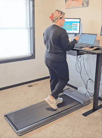 Person walks on a treadmill in an office, working on a laptop at a standing desk with two monitors nearby
