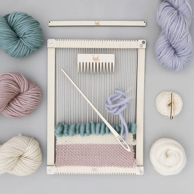 A weaving loom kit featuring partly weaved fabric with balls of yarn and a weaving tool, suitable for crafting and DIY home decor projects