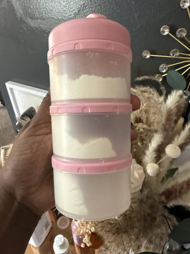 Hand holding a stackable instrumentality  with pinkish  lids, each   conception  filled with powder, successful  a location  mounting  with decorative items successful  the background