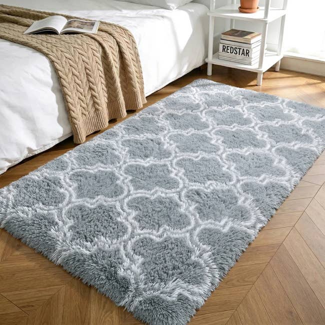 A plush patterned rug beside a bed with a knit blanket, and a small shelf with books and decor in a cozy bedroom setting
