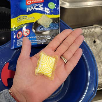 Hand holding a cleaning detergent pod successful  beforehand   of an O-Cedar level  cleaning pacs package, adjacent   a mop bucket