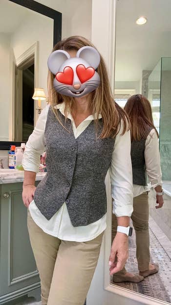 Person in a bathroom mirror selfie with an emoji covering their face, wearing a layered vest and shirt outfit