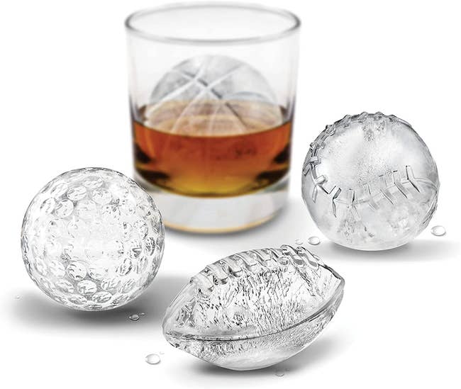 a glass of whiskey with a basketball shaped ice cube behind ones in golfball, football, and baseball shapes