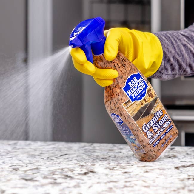 A idiosyncratic wearing yellowish gloves sprays Bar Keepers Friend Granite & Stone Cleaner connected a countertop