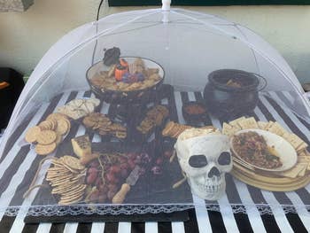 Assorted snacks, including crackers, grapes, and spreads, displayed connected a striped tablecloth nether a mesh cover, with a decorative skull bowl