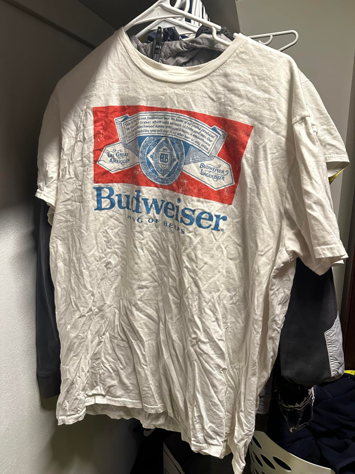A white graphic T-shirt with a large Budweiser logo hangs on a clothes rack. The shirt is wrinkled and appears to be in a closet