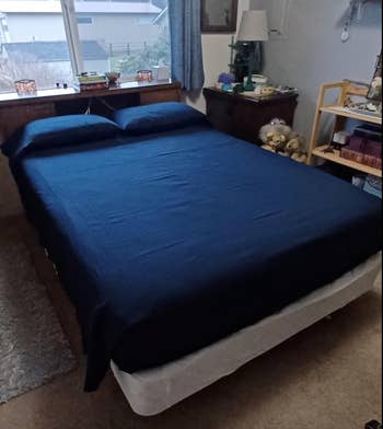A neatly made bed with a blue comforter, set in a bedroom with various furnishings and stuffed animals on a shelf