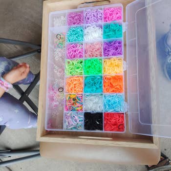 Box of colorful elastic bands and beads organized in compartments, next to a child's legs in star-patterned pants on a chair