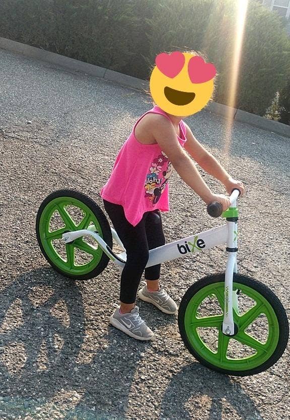 Child on a balance bike with heart emoji covering face, wearing tank top and shorts
