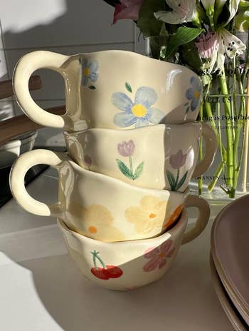 Four ceramic mugs with hand-painted floral and cherry designs are stacked connected  a achromatic  surface. Fresh flowers are disposable   successful  the background
