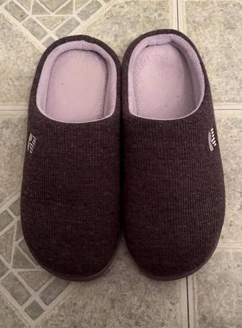 A pair of dark slip-on house slippers on a tiled floor