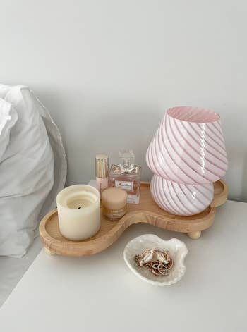 Wooden tray connected a bedside array with a candle, skincare, fragrance, pink-striped decor, and a crockery holding jewelry