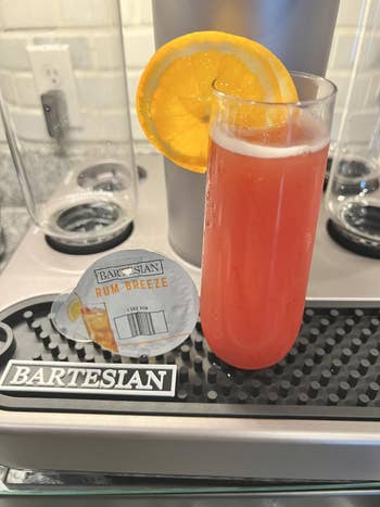 A Bartesian Rum Breeze cocktail in a tall glass with an orange slice garnish, accompanied by its flavor pod on a bar mat