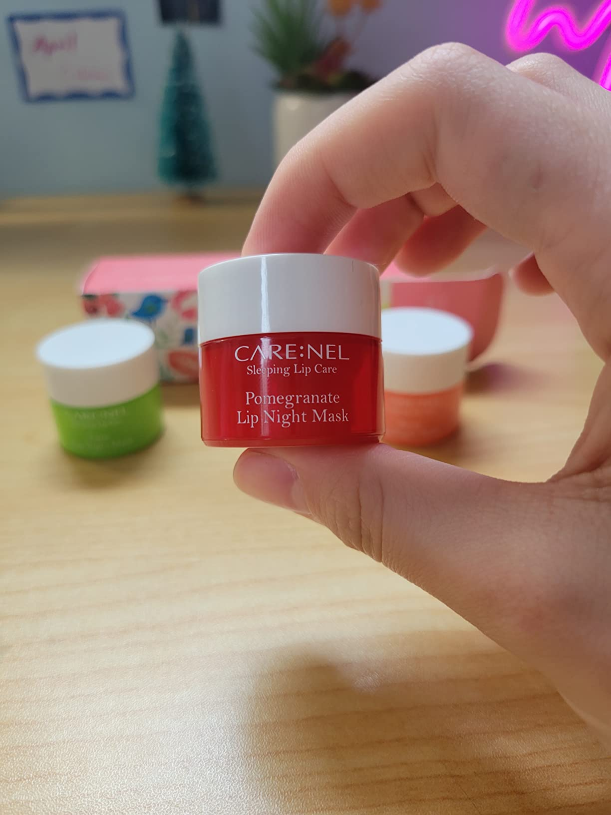 Hand holding a jar of CARE:NEL Pomegranate Lip Night Mask with other skincare products blurred in the background on a wooden surface