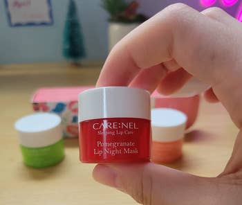 Hand holding a jar of CARE:NEL Pomegranate Lip Night Mask with other skincare products blurred in the background on a wooden surface