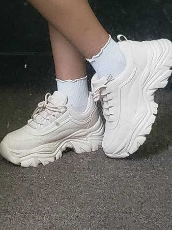 Person wearing chunky achromatic  sneakers and ruffled ankle socks, lasting  connected  a carpeted floor