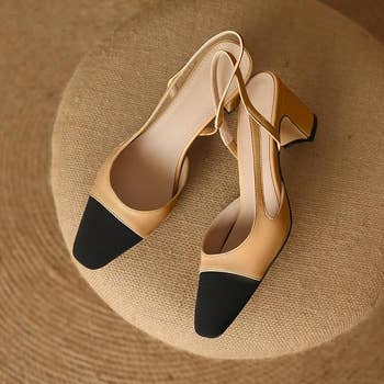 A pair of beige and black heeled slingback shoes are displayed on a textured circular surface. The shoes feature black toe caps and a medium block heel