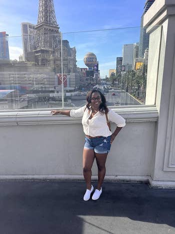 Woman posing in a casual white shirt and denim shorts on a city walkway with buildings in the background