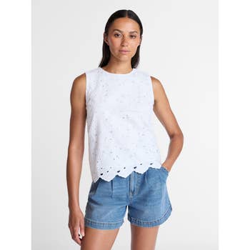 Person wearing a sleeveless eyelet top and denim shorts, posing with hands in pockets against a plain backdrop