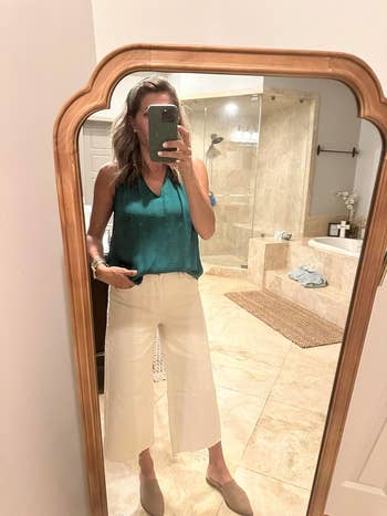 A reviewer takes bathroom mirror selfie wearing a sleeveless top and wide-leg pants. Room includes shower and bathtub in background