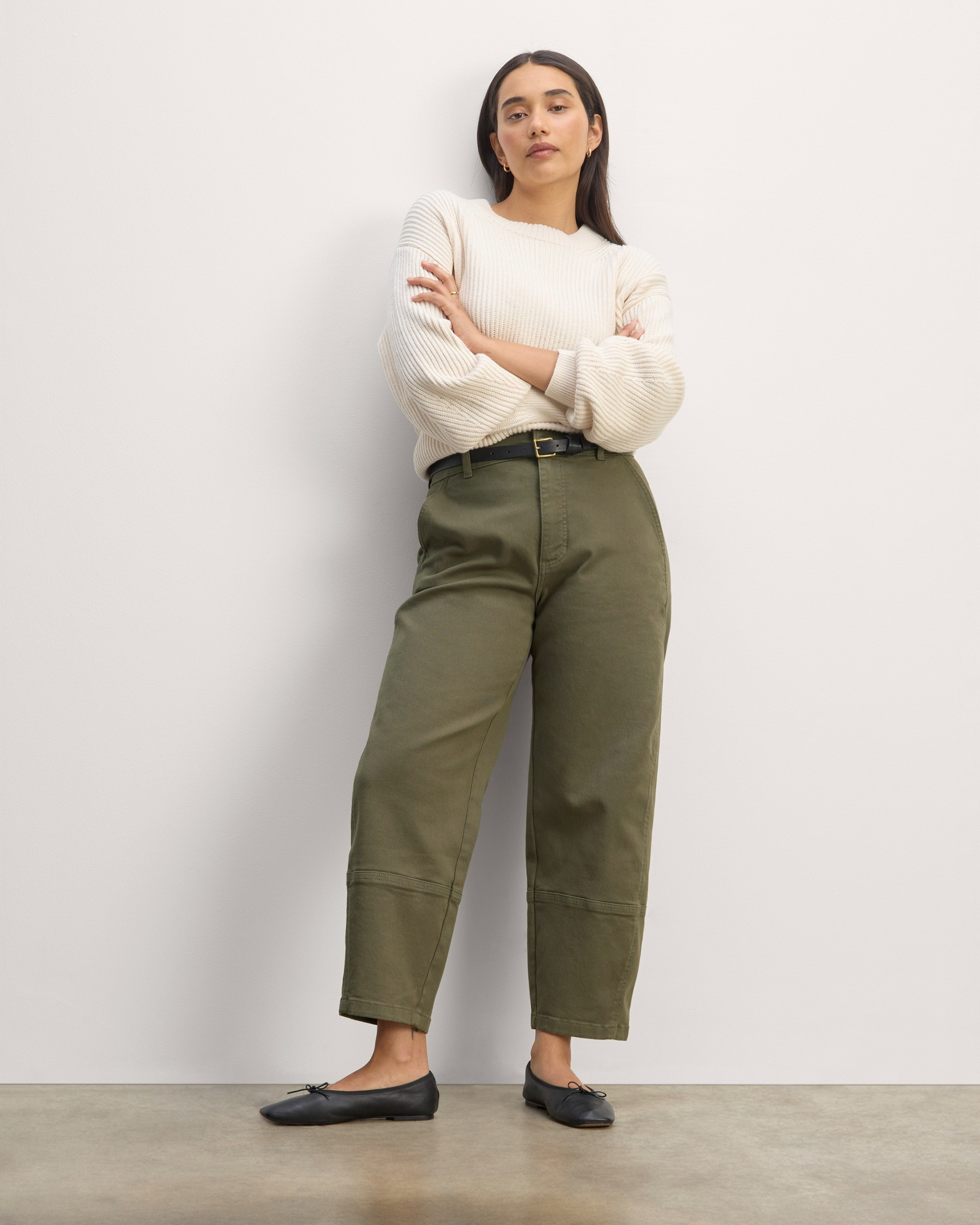 A model in a cream sweater with rolled sleeves, olive green high-waisted pants, and black loafers, standing against a plain background