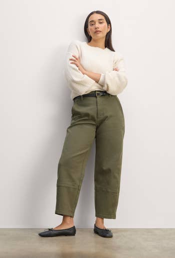 A model in a cream sweater with rolled sleeves, olive green high-waisted pants, and black loafers, standing against a plain background