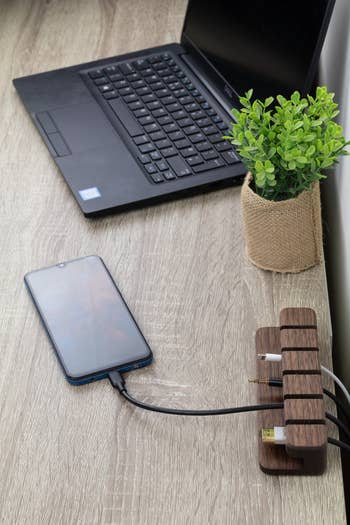 The dark wood organizer holding four different cords on a wood desk charging a phone near a laptop