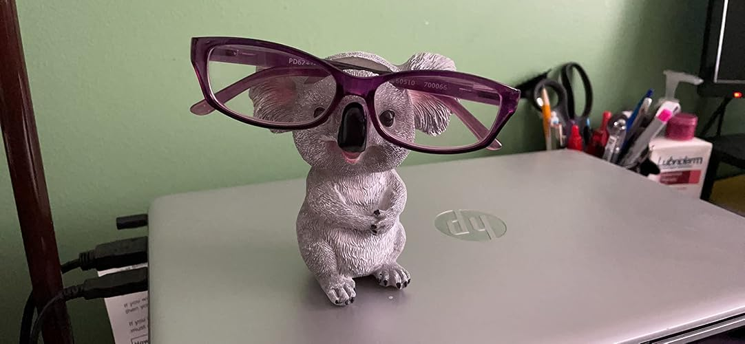 Koala-shaped eyeglass holder sits on a laptop, holding a pair of glasses under its nose. Office supplies are visible in the background