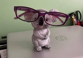 Koala-shaped eyeglass holder sits on a laptop, holding a pair of glasses under its nose. Office supplies are visible in the background