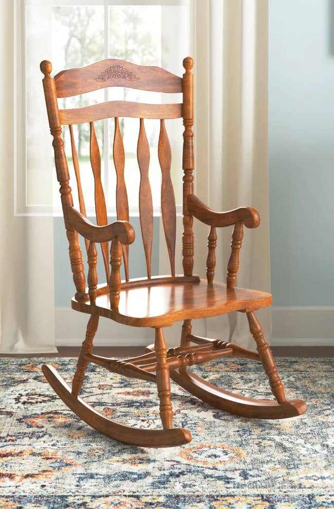 Brown solid wood rocking chair with curved slatted backing and armrests, and a floral pattern engraved on top, sitting on a blue, yellow, and white vintage carpet