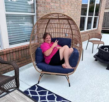 Person sitting successful a wicker ovum seat connected a patio, giving a bid sign. The setup includes different wicker furnishings and a patterned outdoor rug