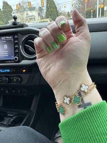 Person wearing a golden  bracelet with clover-shaped charms, displaying green-themed nail art, sitting successful  a car   interior