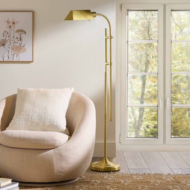 Cozy living room corner with a plush armchair, elegant brass floor lamp, and framed botanical art beside a sunlit window