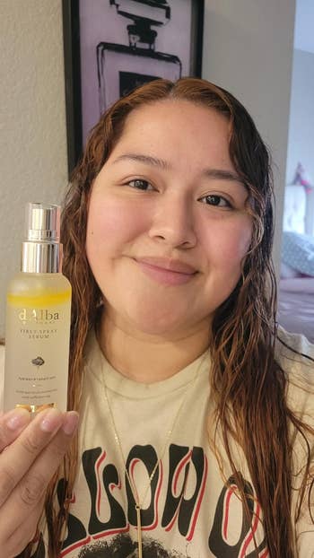 Person smiling, holding a d'Alba mist spray bottle. They person  wavy hairsbreadth  and deterioration  a graphic T-shirt. Background shows a framed representation  connected  the wall