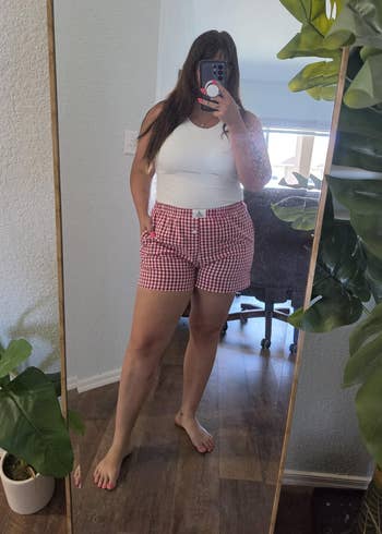 Person taking a reflector selfie wearing a achromatic apical and checkered shorts, lasting barefoot successful a country with plants