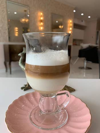 Reviewer photo of a layered latte in a clear glass on a pink saucer, with a blurred salon interior in the background