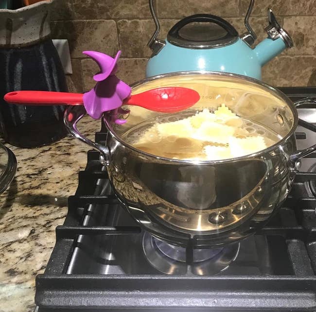 Cooking pot on stove with pasta, red spoon, and a purple witch spoon holder atop the handle