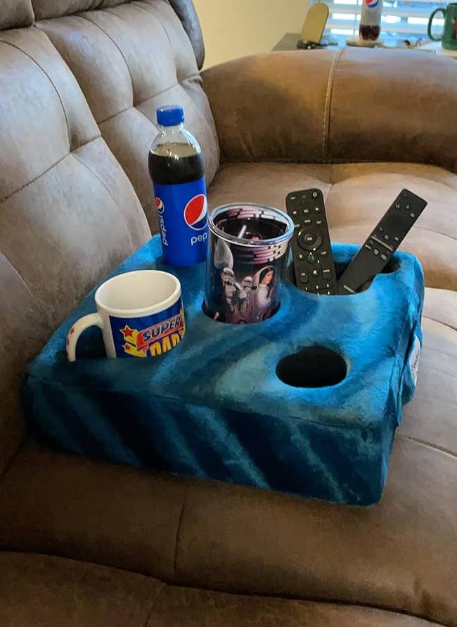 A Super Mario mug, Pepsi bottle, patterned cup, and remote controls on a blue couch arm tray